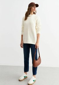 Maglione crema a maglia grossa, jeans scuri, sneakers bianche con accenti verdi, borsa marrone. Outfits informale con materiali testurizzati e vestibilità rilassata.