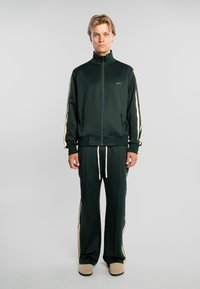 Joven con cabello rubio que lleva un chándal verde oscuro con rayas crema y zapatos beige, de pie frente a un fondo gris liso.