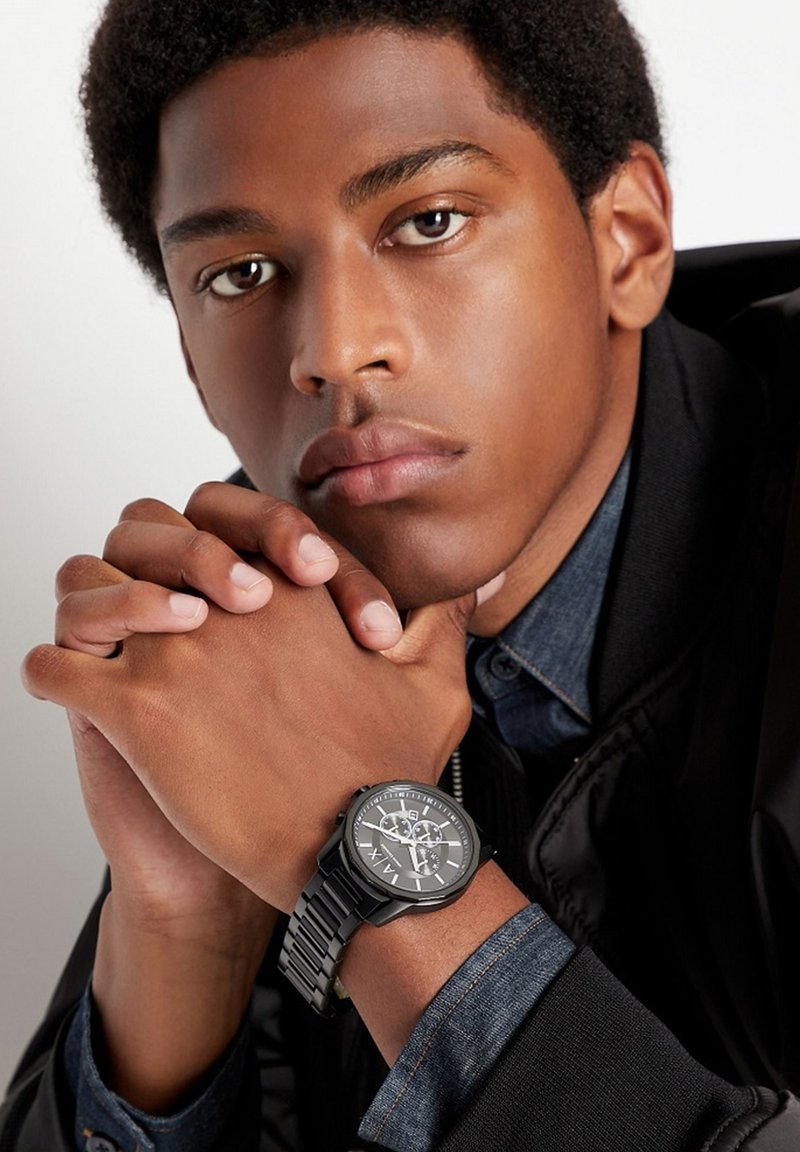 Schwarze analoge Uhr mit detailiertem Zifferblatt, silbernen Akzenten und einem strukturierten Gummiband, getragen an einem Handgelenk mit gefalteten Händen.