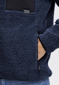 Pullover in fleece blu navy con tessuto testurizzato, una toppa nera sul petto e una tasca laterale con zip. Caratterizzato da un materiale morbido e accogliente e da un design casual.