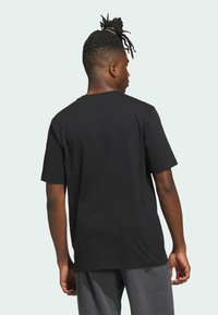 Černé bavlněné tričko s krátkými rukávy, kulatým výstřihem a volným střihem, z pohledu zezadu. V kombinaci se šedými tepláky.