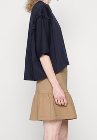 Una donna vestita con una blusa nera ampia con rouches sulle maniche e una gonna beige svasata, mostrata di lato contro uno sfondo neutro.