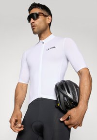 Maillot de cyclisme blanc à manches courtes avec une fermeture éclair à l'avant, logo noir ; associé à un short de cyclisme noir et un casque noir tenu dans une main.