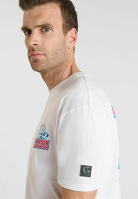 Man met kort bruin haar die een wit T-shirt draagt met een rood en blauwe graphic "Attention Angry Boss" en een zwart logo patch op de mouw.