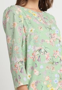 Blommig skjorta i mjukt grönt tyg med V-ringning och spetsdetaljer. Designen inkluderar flerfärgade blommor och en avslappnad passform.