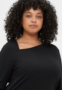 Μαύρο μπλουζάκι με μακριά μανίκια και μοναδική ασύμμετρη λαιμόκοψη. Λείο ύφασμα, εφαρμοστή γραμμή και υφή στη ραφή του ώμου.