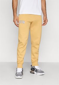 Άνδρας φορώντας μπεζ φόρμες με κείμενο "BOSTON" στον αριστερό μηρό και μαύρα-άσπρα αθλητικά παπούτσια, στέκεται μπροστά σε ένα ουδέτερο φόντο.