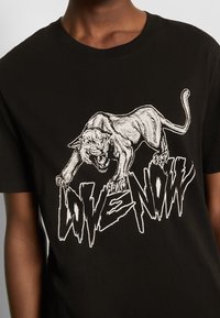 Czarny t-shirt z białą grafiką warczącego pantera nad stylizowanym napisem „LIVE NOW” w postrzępionych literach.
