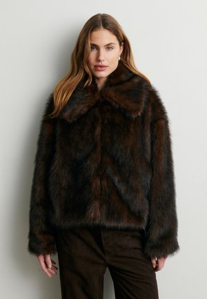 Jeune femme aux longs cheveux portant une veste en fausse fourrure marron foncé et un pantalon sombre assorti, se tenant contre un mur clair uni.