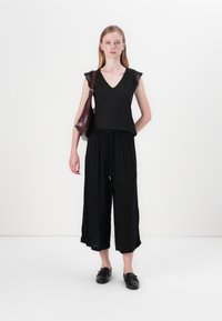 Juodas be rankovių topas su nėriniu, derinamas su laisvomis juodomis culotėmis. Modelis laiko bordo spalvos petinę rankinę ir vilki juodas slip-on tipo batus.