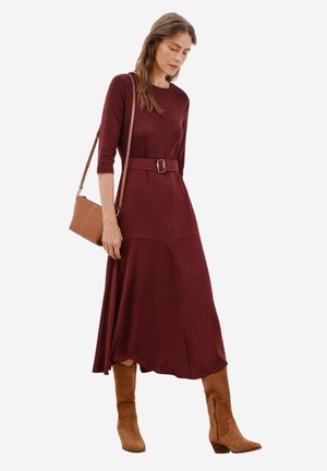 Bordeauxfarbenes Strickkleid mit Dreiviertelärmeln, einem Gürtel in der Taille und einem fließenden Rock. Kombiniert mit beigen Stiefeletten und einer Umhängetasche.