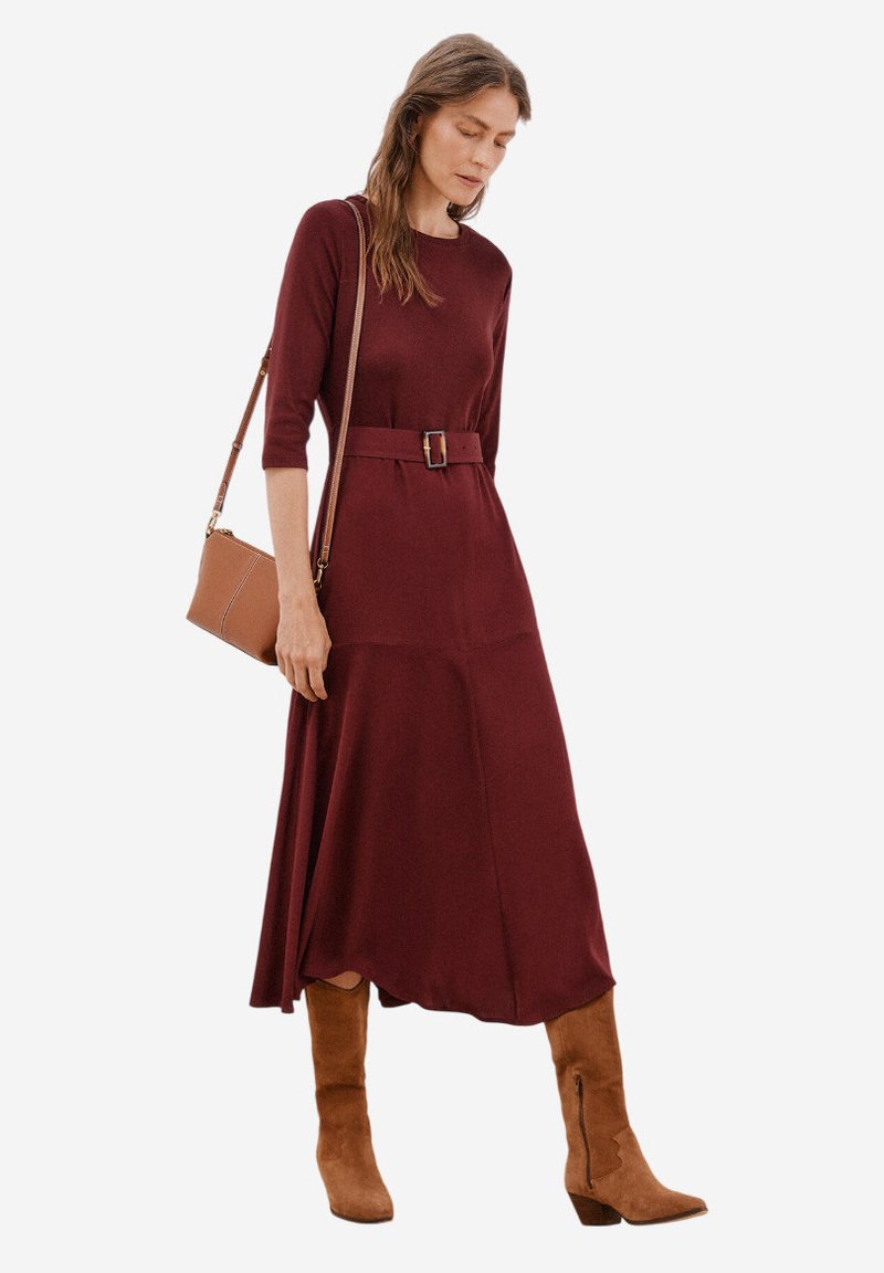 Bordeauxfarbenes Strickkleid mit Dreiviertelärmeln, einem Gürtel in der Taille und einem fließenden Rock. Kombiniert mit beigen Stiefeletten und einer Umhängetasche.