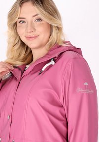 Giacca impermeabile rosa con cappuccio, dotata di cordini bianchi e bottoni a pressione. Logo sulla spalla sinistra, texture liscia.