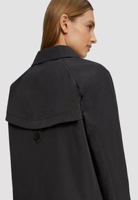 Zwarte trenchcoat met een spitse kraag, achterkant met klepdetail en een knoopsluiting. Gemaakt van soepel, textuurstof.