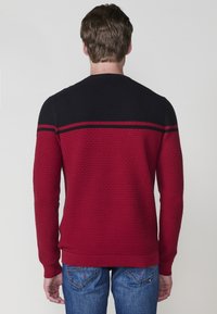 Pullover texture rosso e nero con scollo rotondo, polsini a coste e una striscia orizzontale sulla schiena. Indossato con jeans blu.
