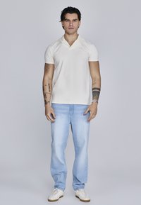 Lys beige polo-shirt med krave, parret med lyseblå jeans. Modellen har hvide sneakers på. Simpelt design og tætsiddende silhuet.