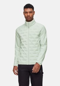 Jasnozielona, ocieplana kurtka z pikowanym wzorem, wysoki kołnierz, pełny zamek błyskawiczny i elastyczne mankiety. Noszona z czarnymi spodniami.