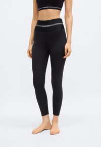 Donna che indossa leggings a vita alta neri Calvin Klein e reggiseno sportivo abbinato, in piedi scalza su uno sfondo bianco semplice.