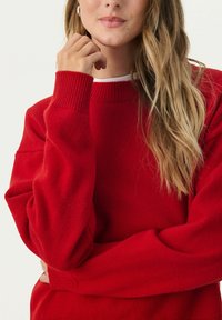 Red knit sweater with ribbed cuffs and neckline. Soft texture, relaxed fit. Model poses with one arm folded and hand near her face.