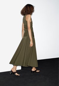 Olijfgroene, mouwloze maxi-jurk met een getailleerde taille en een vloeiende rok, gecombineerd met open teentjes zwarte blokhak sandalen op een zwart oppervlak.