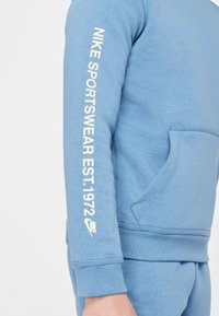 Blue cotton sweatshirt featuring a kangaroo pocket, ribbed cuffs, and white text on the left sleeve: "NIKE SPORTSWEAR EST. 1972" with logo.