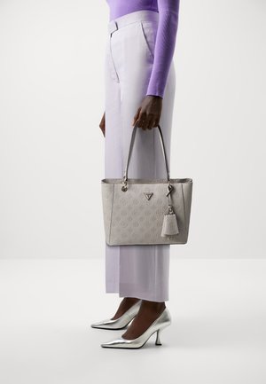 Personne en haut lilas et pantalon clair tenant un sac fourre-tout beige embossé, portant des chaussures à talons hauts métalliques argentées sur un fond blanc.