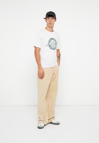 Hvit T-skjorte med korte ermer og grønn grafikk; kombinert med løse beige bukser. Svart cap; grå og hvite joggesko. Enkelt, uformelt antrekk.