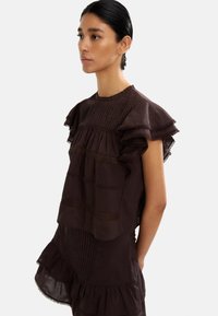 Brązowa bluzka z tkaniny z falbaniastymi rękawami, zdobiona detaliami w postaci zakładek oraz koronkowymi akcentami. Projekt wyróżnia się warstwowymi teksturami i luźnym krojem.