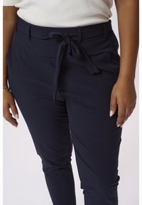 Mørkeblå bukser med en glat overflade, der har et bundet bælte i taljen og sidelommer for funktionalitet.