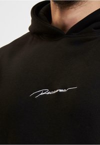Zwarte hoodie van zacht materiaal met een witte geborduurde logo op de borst. De halslijn heeft een standaard capuchonontwerp.