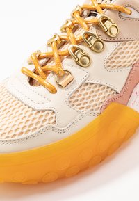 Leichter Sneaker mit einem Obermaterial aus Mesh in Beige, pinken Akzenten, einem gelben Schnürsystem und einer durchsichtigen, orangefarbenen Gummisohle mit strukturiertem Muster.