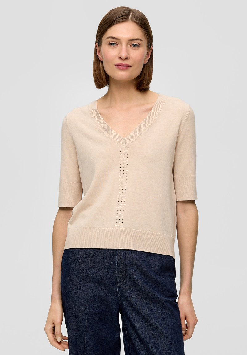 Beige Kurzarm-V-Ausschnitt-Pullover mit strukturiertem Muster an der Vorderseite und einem gerippten Saum. Getragen mit dunkelblauen Jeans.