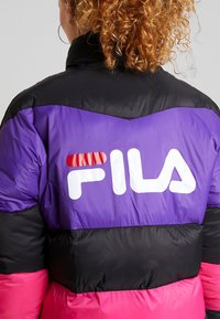 Personne aux cheveux bouclés portant une doudoune FILA noire, violet et rose avec le logo dans le dos, de dos à la caméra.