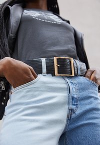Cinto texturizado preto com uma fivela quadrada dourada usado sobre jeans de denim azul claro. O cinto apresenta um design mais resistente e trançado.