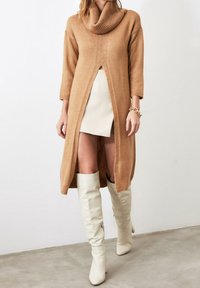 Robe tricotée couleur fauve avec un col bénitier, manches 3/4 et une fente à l'avant, complétée par des cuissardes blanches. Texture lisse et coupe décontractée.