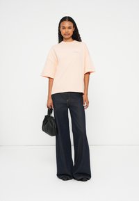 T-shirt oversized rosa claro com mangas curtas, combinada com jeans de perna larga azul escuro e uma bolsa preta franzida. Design e tecido simples.