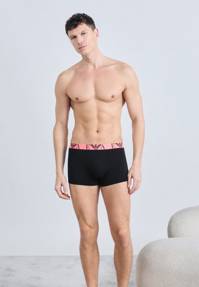 Homme torse nu debout avec une expression neutre, portant un boxer noir avec une ceinture rose affichant le logo de la marque.