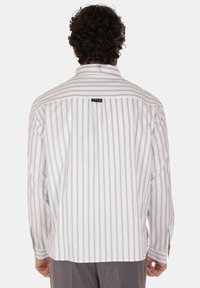 Camicetta bianca a strisce con linee verticali viola, maniche lunghe e yoke sul retro. Presenta un piccolo etichetta del marchio al collo. Vestibilità sartoriale.