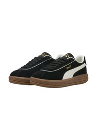 Zapatillas de ante negro con una franja lateral blanca, suela de goma marrón y una marca dorada en la lengüeta y el lateral. Diseño con cordones, estilo bajo.