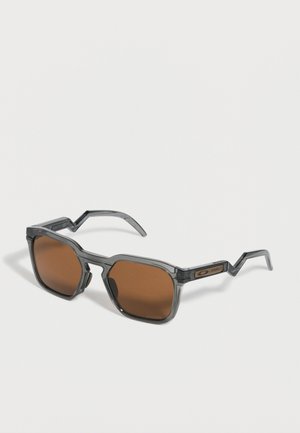 Lunettes de soleil rectangulaires avec une monture translucide gris fumé et des verres marron, avec des branches angulaires en forme de zigzag.