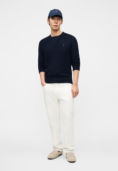 Πλεκτό πουλόβερ ναυτικού μπλε με στρογγυλή λαιμόκοψη, κρεμ παντελόνια και ανοιχτό μπεζ μοκασίνια. Καπέλο μπέιζμπολ με κόκκινο λογότυπο ολοκληρώνει το σύνολο.