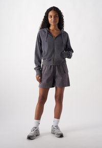 Grau zip-up Hoodie mit Zugbändern, kombiniert mit passenden grauen Shorts mit Taschen. Modell trägt graue Sneakers und weiße Crew-Socken.