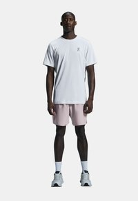 Weißes T-Shirt mit kurzen Ärmeln und Logo, kombiniert mit hellrosa Shorts. Das Modell trägt auch weiße Socken und graue Sportschuhe.