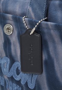 Blauwe stoffen boodschappentas met een gestructureerde weefsel, zilveren hardware en een zwart leren label met een geëmbosseerd logo, bevestigd met een metalen ketting.