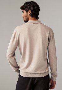 Ljusbeige tröja med långa ärmar, en subtil textur och ribbad nederdel. Har en krage och en avslappnad passform, sedd från baksidan.