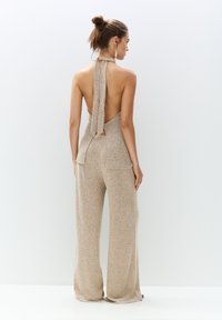 Back view of a halter-neck jumpsuit in a beige, textured fabric with a wide-leg design and a tied detail at the back.