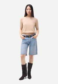 Pull beige en maille, short en denim bleu clair taille haute avec ourlet effiloché, ceinture marron, et bottes noires montantes à bout pointu.