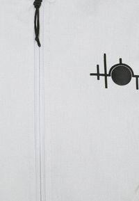 White zip-up jacket made of textured fabric featuring a black graphic logo design on the left side. Includes adjustable drawstring at the collar.