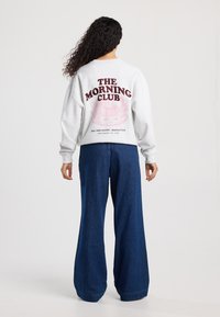 Grijze sweatshirt met ronde halslijn en oversized pasvorm, met "THE MORNING CLUB" in donker kastanjebruin en een grafisch ontwerp op de achterkant.