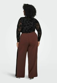 Žena s kovrčavom kosom nosi crni cvjetni prozirni top dugih rukava i smešu široke nogavice smešnih hlača s crnim bočnim trakama, okrenuta je leđima.